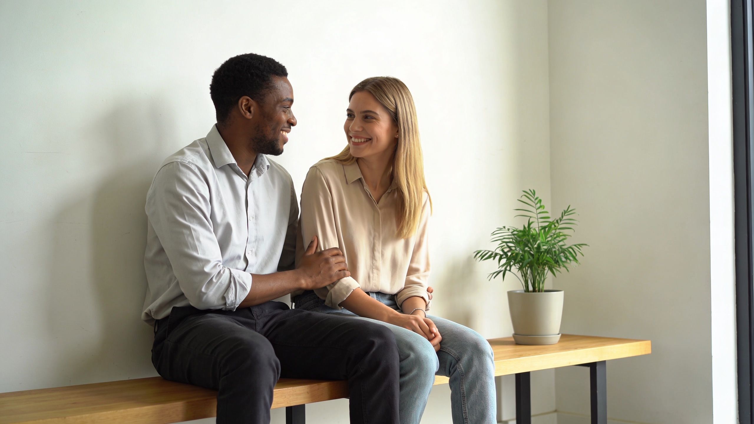 A happy multiracial couple sitting on a wooden bench in a bright office environment during a break.
