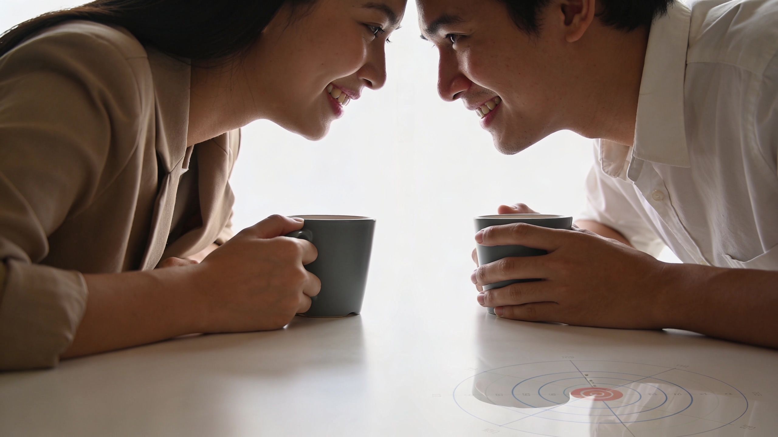 A happy couple sitting at a table with coffee mugs, smiling at each other in a bright room.