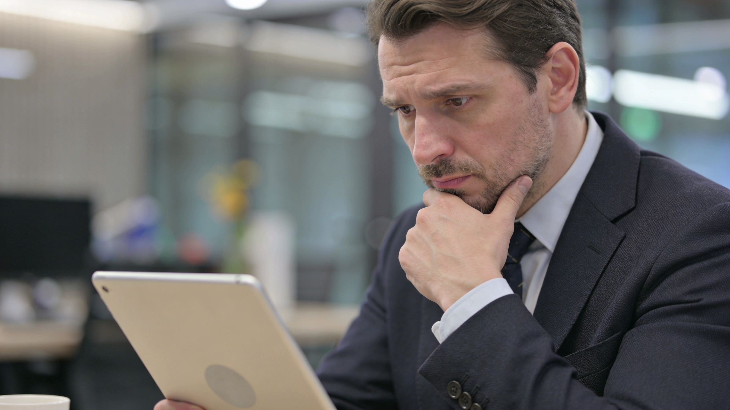 A professional businessman in a suit intently examines data on a digital tablet while sitting in an office.