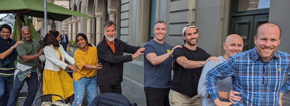 A diverse group of smiling people outdoors in a city, forming a human chain with hands on shoulders.