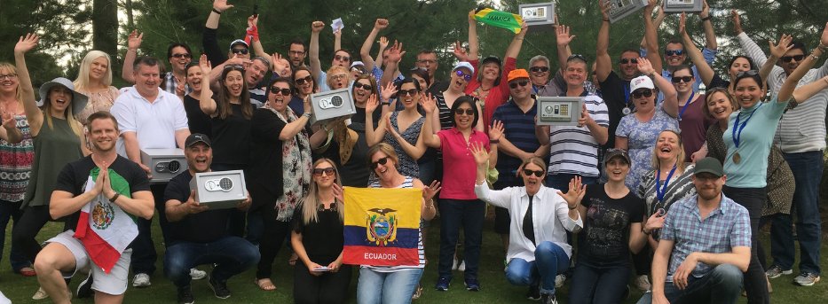 A large group of smiling people outdoors, some holding flags and safes, celebrating a team-building event.