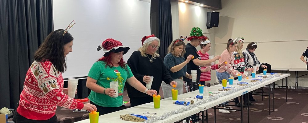 Women in festive wear play a game with ping-pong balls and cups at an end-of-year event.