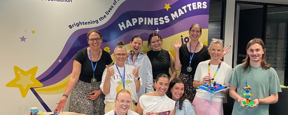 A group of happy colleagues pose with Lego creations in front of a 'Happiness Matters' mural.