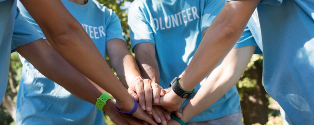 Group of volunteers in blue shirts stack hands together, symbolizing teamwork and unity outdoors.