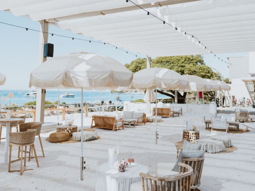 Outdoor beach club with white sand, fringed umbrellas, wooden seating, and clear blue water.