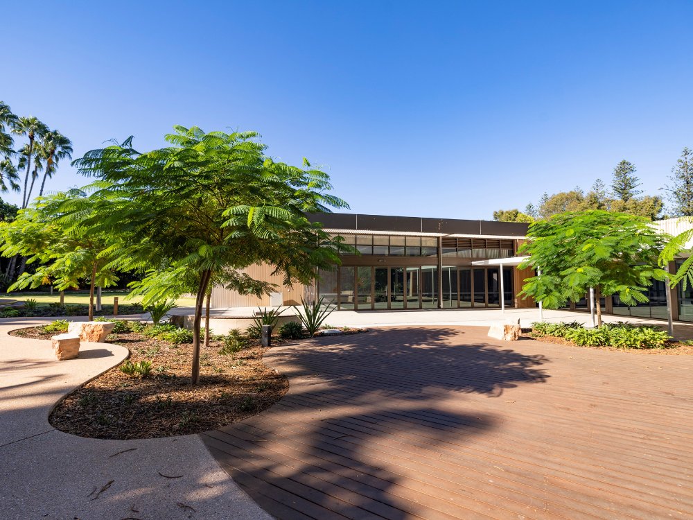 Modern building with large windows, surrounded by lush green trees, garden beds, and a spacious outdoor deck.