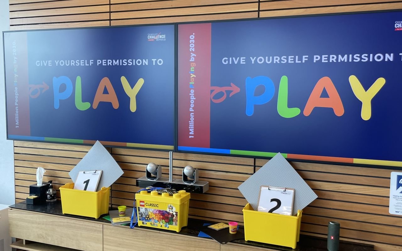 Two digital screens display the message Give Yourself Permission To Play above two yellow activity bins.