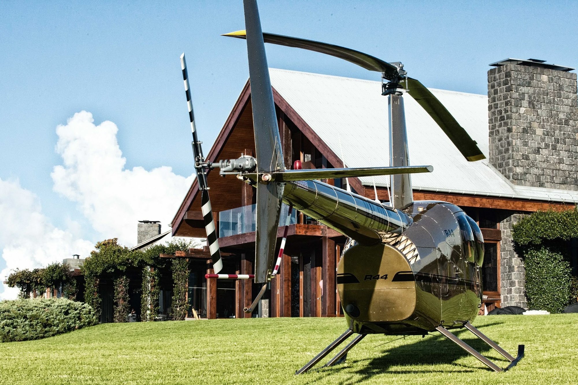 A Robinson R44 helicopter stands on a lush green lawn, with a large house in the background.