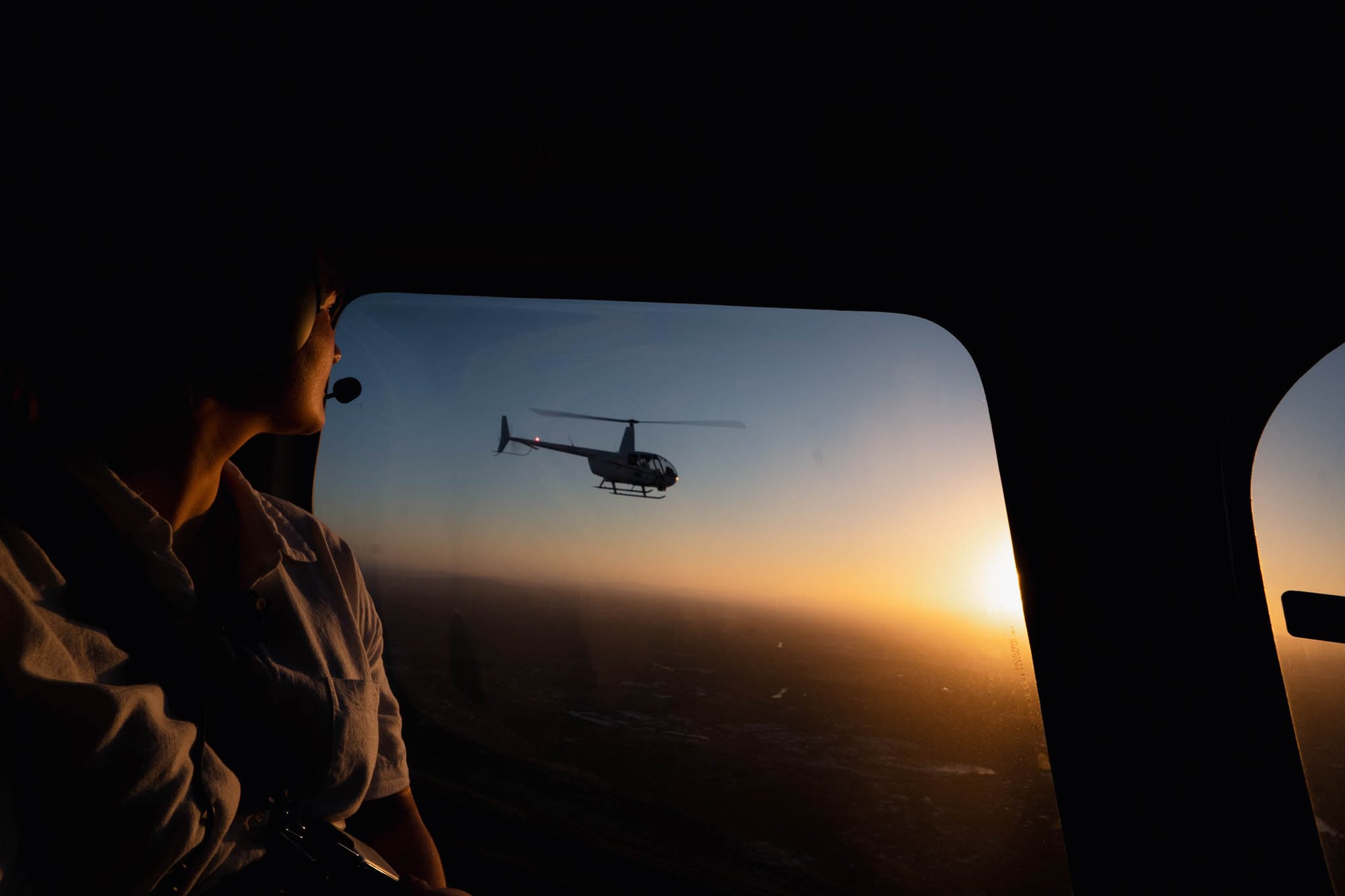 A pilot inside a helicopter views another helicopter flying against a beautiful sunset sky.
